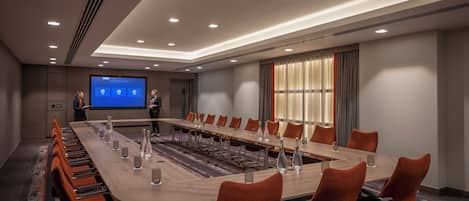Un salón de conferencias con una larga mesa, sillas anaranjadas, una pantalla de proyector y un amplio ventanal con cortinas.