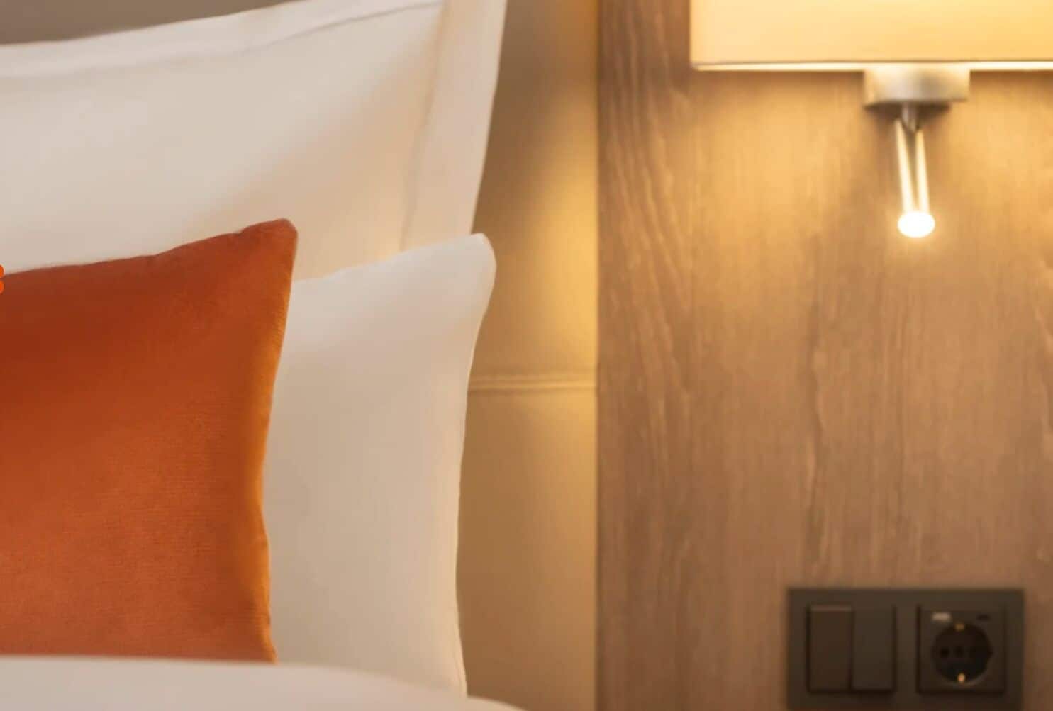 A bed with white and orange pillows, a wall-mounted light, and a power outlet.