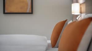 A neatly made bed with white and orange pillows, a framed artwork on the wall, and a bedside lamp.