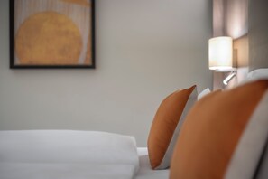 Een opgemaakt bed met witte en oranje kussens, een ingelijst kunstwerk aan de muur en een bedlampje.