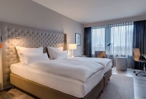 Una habitación de hotel con una cama grande, cabecera, escritorio, silla, lámpara y ventana con cortinas.