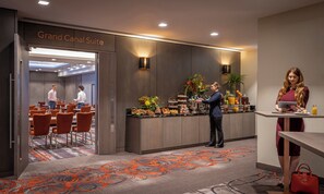 Een hotel lobby met een receptiebalie, een bordje ‘Grand Canal Suite’, een buffet met diverse gerechten en een zithoek met oranje stoelen.