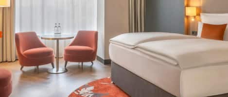 Une chambre d’hôtel avec un lit, deux fauteuils, une petite table et une fenêtre avec des rideaux.