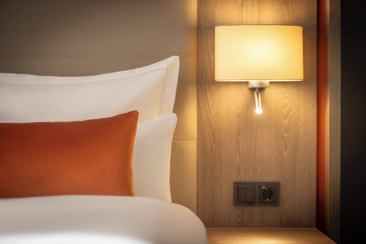 A neatly made bed with white and orange pillows, a wall-mounted lamp, and a power outlet.