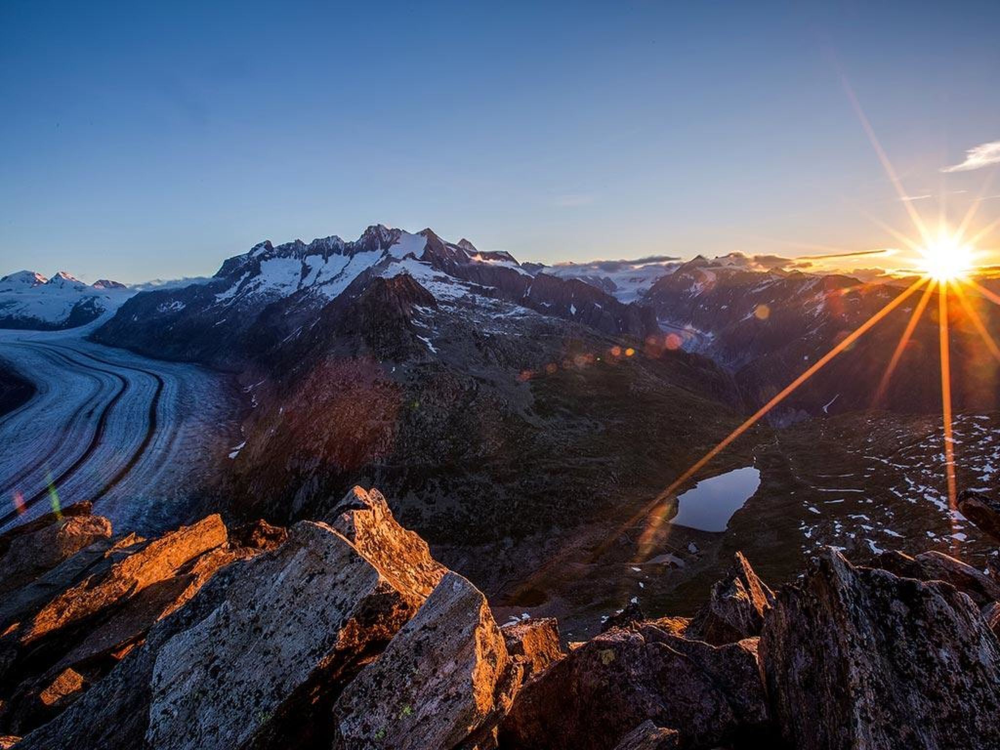 Paisatge de muntanyes amb un glaçer, terreny rocos i un sol ixent.