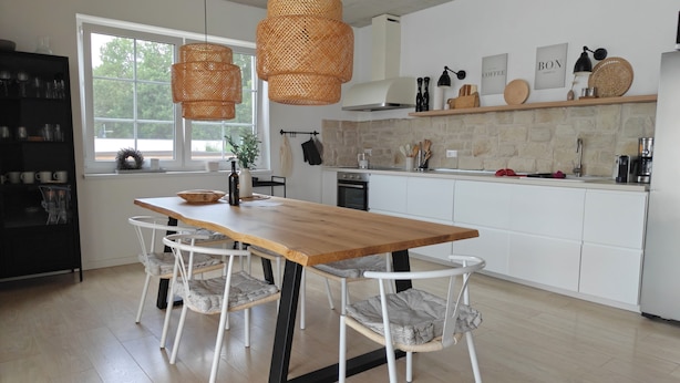 Eine moderne Küche mit einem Holzesstisch, weißen Stühlen, hängenden Korbtreleuchtern und einer Stein-Spritzschutzleiste.