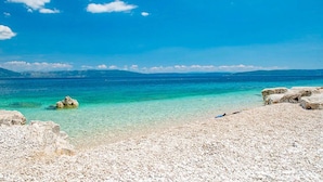 Ein Strand mit klarem türkisblauem Wasser, felsigem Ufer und Bergen in der Ferne unter blauem Himmel.