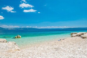 Ein Strand mit klarem türkisblauem Wasser, felsigem Ufer und Bergen in der Ferne unter blauem Himmel.