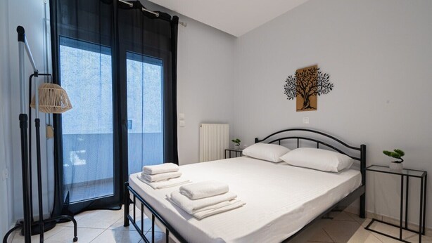 A modern bedroom with a bed, a small bedside table, a radiator, a window with curtains, and a wall-mounted artwork. - V & G Apartment by Rentals Pro - Nea Moudania (Nea Moudania)
