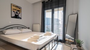A neatly made bed with white linens and towels, a black metal headboard, a large window with dark curtains, a small table with a potted plant, and a mirror on the wall. - V & G Apartment by Rentals Pro - Nea Moudania (Nea Moudania)