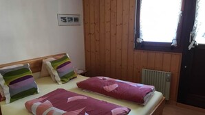 Una cama doble con almohadas a rayas y una manta enrollada, paredes de madera, una ventana con cortinas y un radiador.