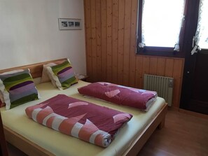 A double bed with striped pillows and a rolled-up blanket, wooden walls, a window with curtains, and a radiator. - Almenrausch 2. OG Süd-West Appartement/Fewo, Dusche, WC by Interhome (Riederalp)