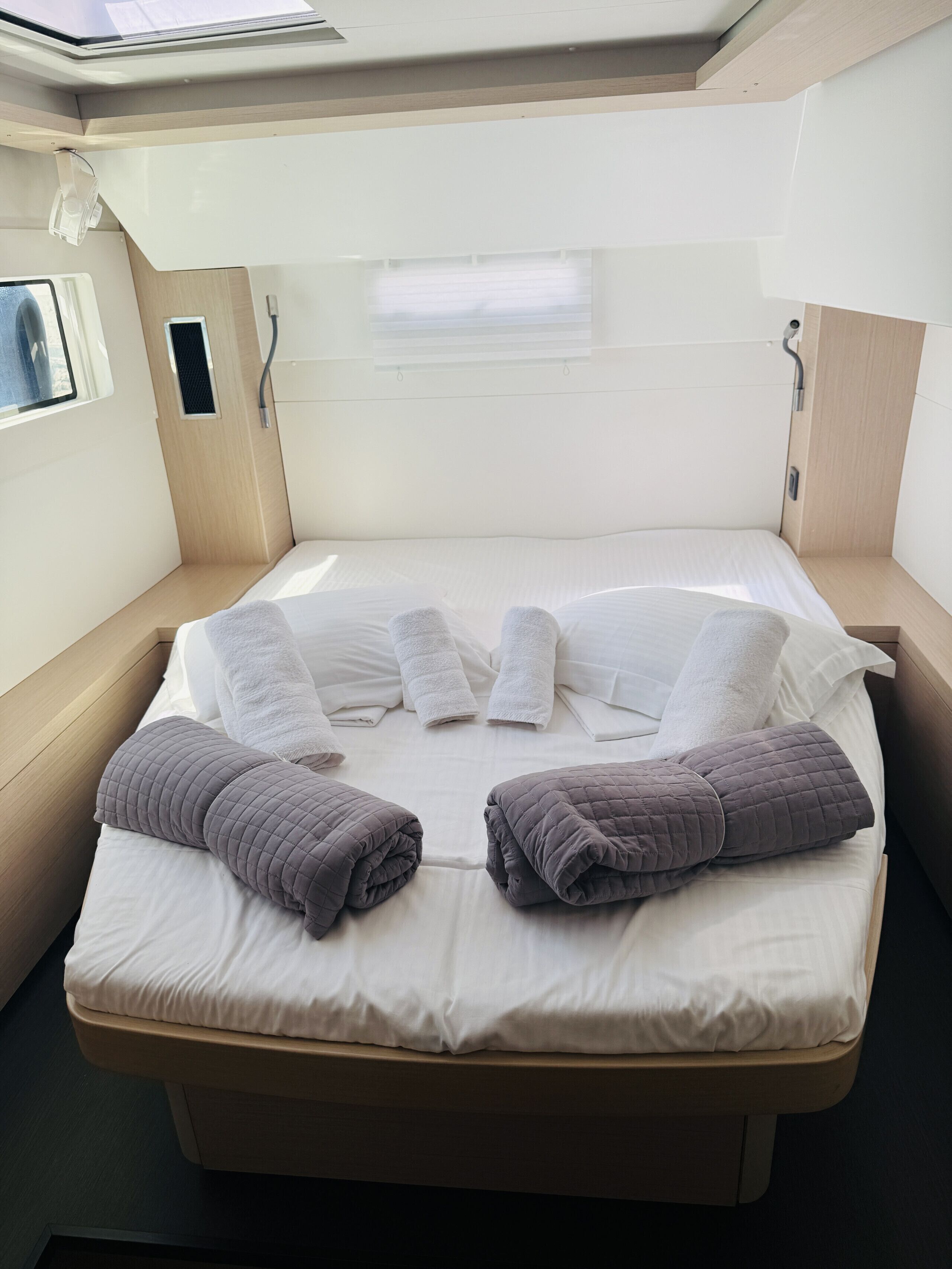 Una cama compacta con ropa blanca y dos mantas enrolladas, una ventana con persianas, y una mesita de noche pequeña.