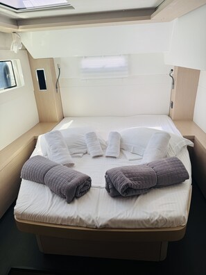 Uma cama compacta com roupa de cama branca e duas mantas enroladas, uma janela com persianas e uma pequena mesa de cabeceira.