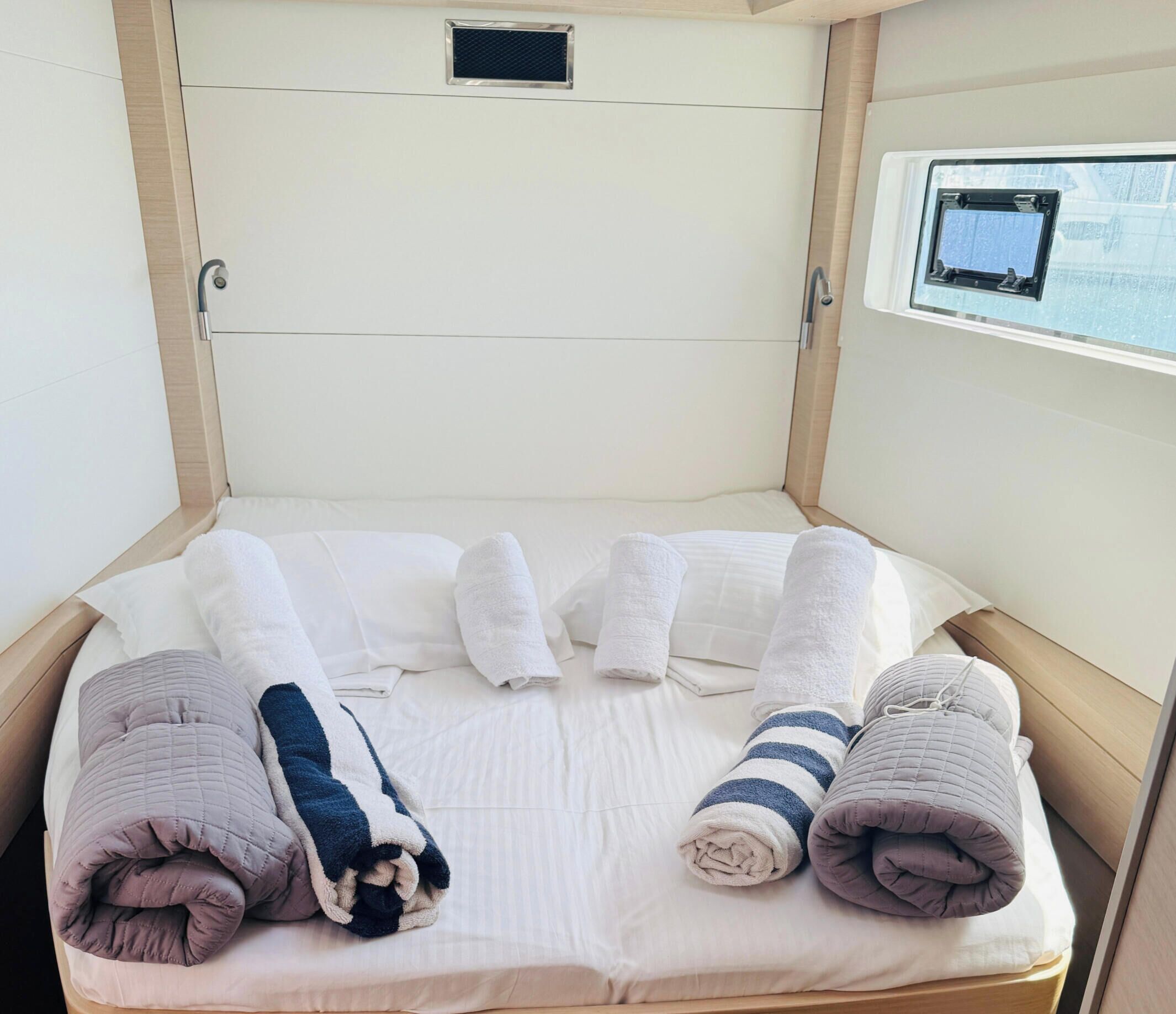 A neatly made bed with rolled towels and a quilted pillow, a window with a view, and a small control panel above the bed.