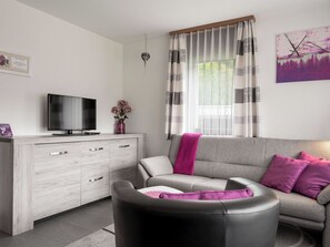 A modern living room with a grey sofa, a black leather chair, a flat-screen TV on a wooden cabinet, and a window with striped curtains.