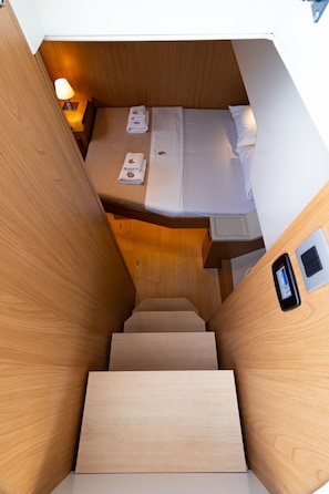 Eine schmale Holztreppe führt zu einem Bett mit Kopfteil, einem Nachttisch mit Lampe und einer Bedienkonsole an der Wand.