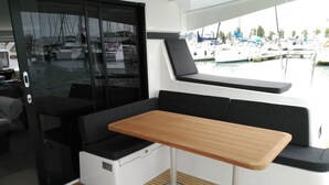 Interior de un barco con una mesa de madera, asientos acolchados negros y vista a un puerto deportivo.