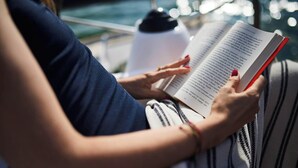 Eine Person liest ein Buch am Fenster mit Blick auf das Wasser.
