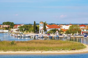 Une ville côtière où des bateaux sont amarrés le long de la rive, un imposant bâtiment blanc surmonté d’un toit rouge, et des immeubles résidentiels en arrière-plan.