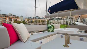 Cubierta de barco con zona de estar acolchada, una mesita y vistas a un puerto deportivo con edificios al fondo.