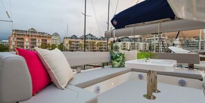 A boat deck with a cushioned seating area, a small table, and a view of a marina with buildings in the background.