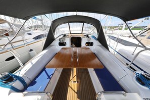 Convés de barco com uma mesa de madeira central, almofadas azuis e vista para outros barcos.