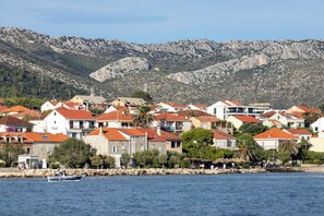 A coastal town with stone buildings, a rocky mountain backdrop, and a body of water with a small boat.