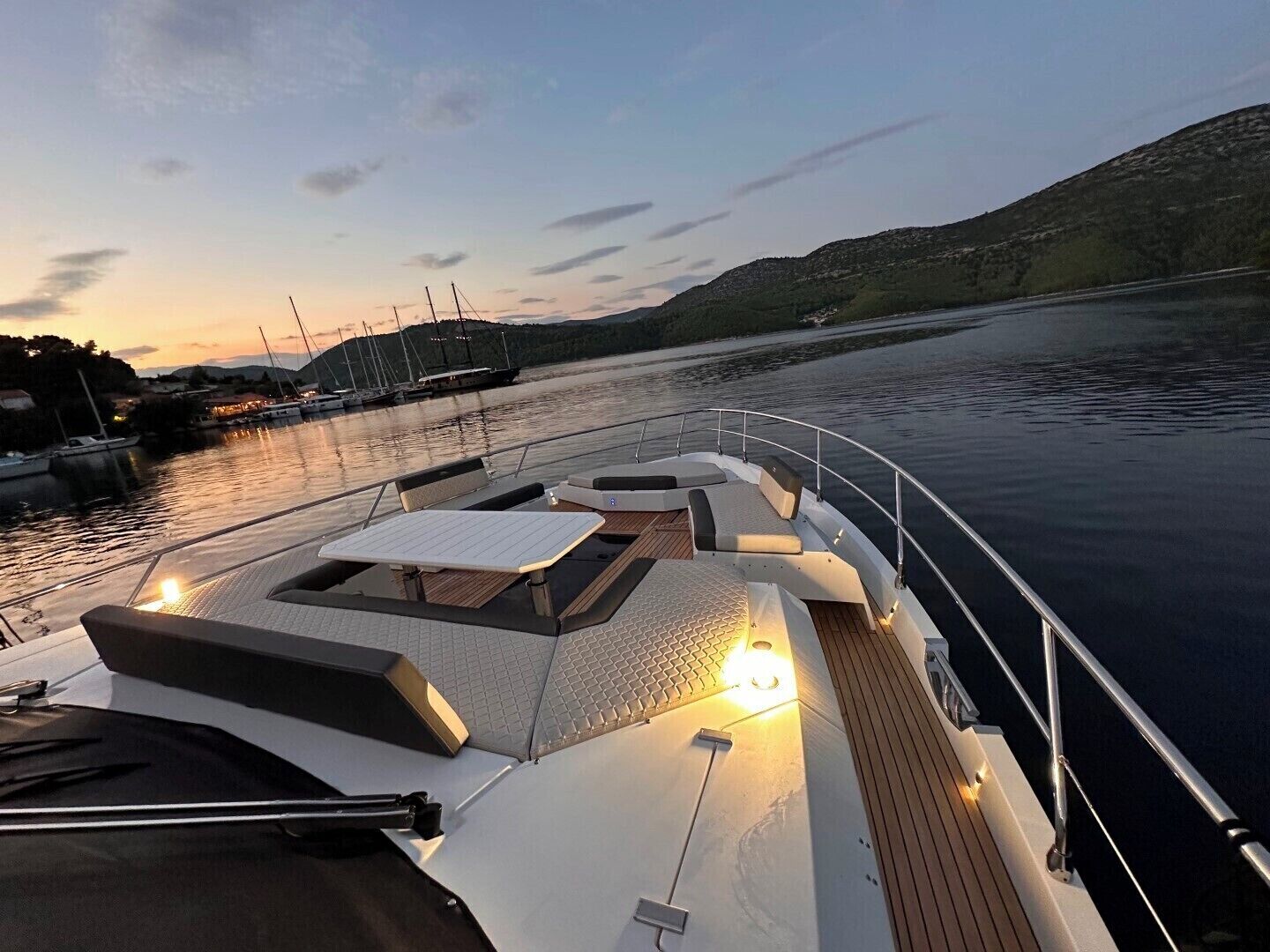 Een dek van een boot met zitplaatsen, een tafel en uitzicht op een jachthaven en heuvels.