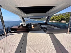 Convés de barco com assentos, uma mesa e vista para o mar e colinas.