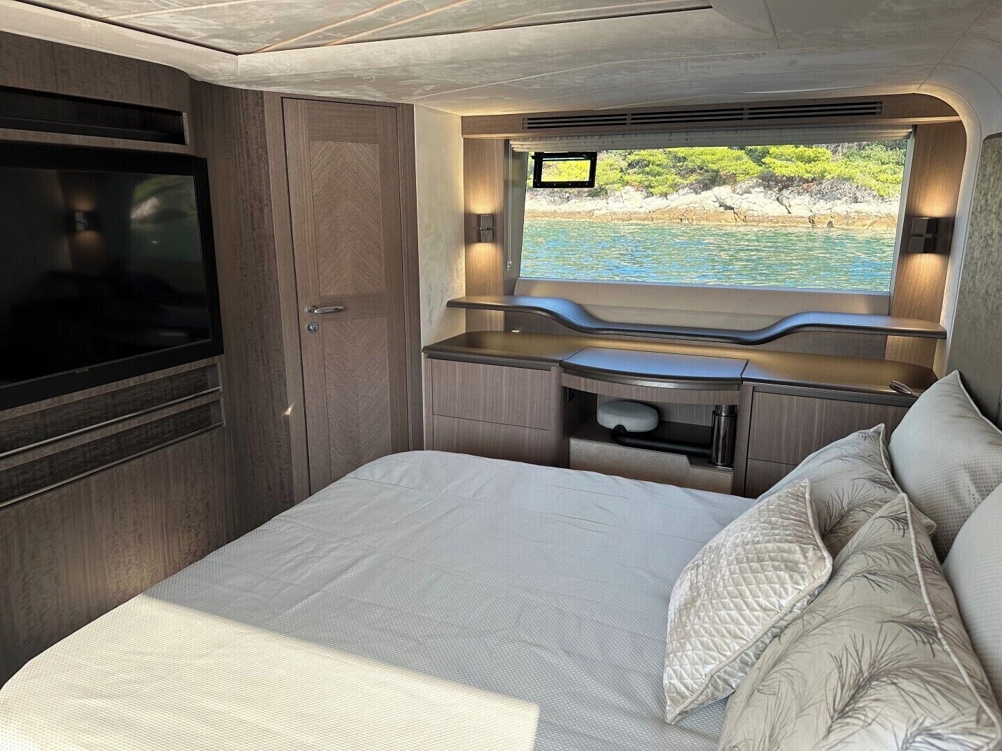 A compact bedroom with a bed, a TV, a window with a view of the sea, and built-in storage.