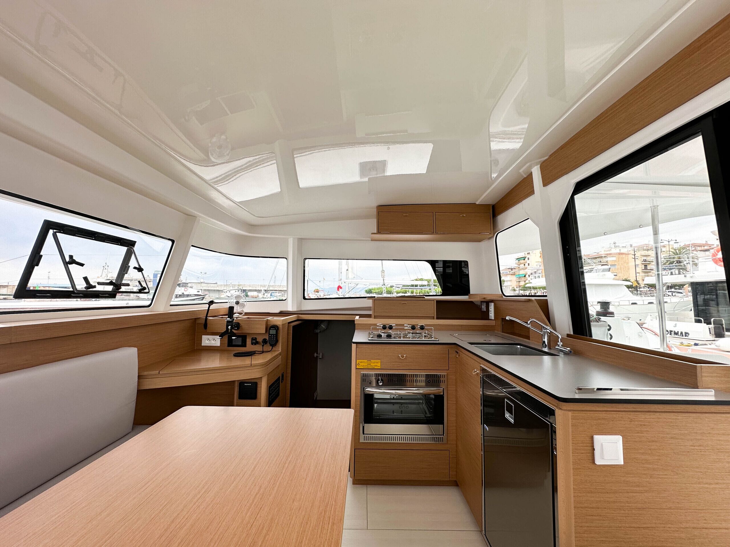 Een kleine kombuis met kookplaat, oven en spoelbak, houten kastjes en grote ramen met uitzicht op de jachthaven.