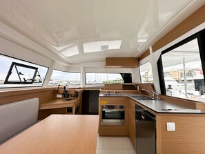 Een kleine kombuis met kookplaat, oven en spoelbak, houten kastjes en grote ramen met uitzicht op de jachthaven.