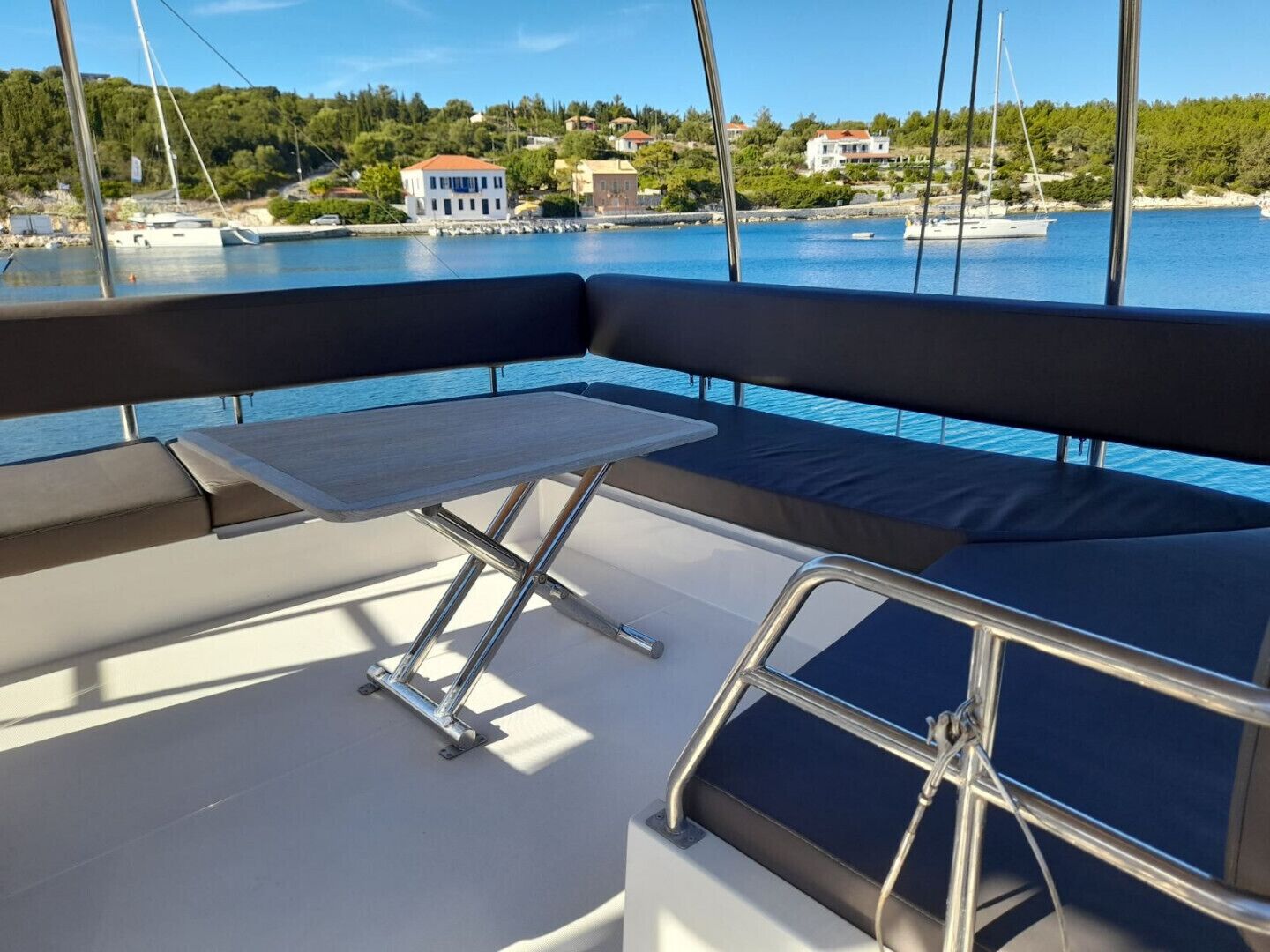 Het dek van een boot met een zitgedeelte met kussens, een kleine tafel en uitzicht op een kustplaats.