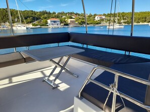 A boat's deck with a cushioned seating area, a small table, and a view of a coastal town. - Sun-Kissed Sailing Adventure in the Ionian Islands (Kontokali)