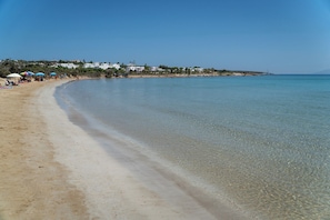 Ein Strand mit klarem Wasser, sandigem Ufer und Gebäuden im Hintergrund.