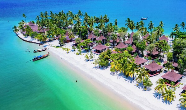 Vista aérea de un complejo turístico en la playa con bungalows sobre el agua, una playa de arena y un mar azul cristalino.