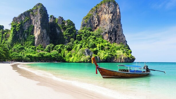 Une plage à l’eau turquoise et limpide, un bateau et une grande formation rocheuse couverte de végétation.