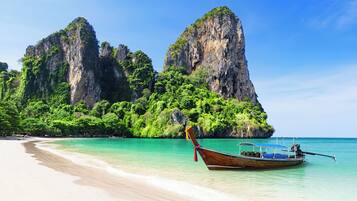 Een strand met helder turquoise water, een boot en een grote rotsformatie met begroeiing.