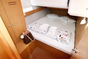 Ein schmales Bett mit weißer Matratze, zwei Kissen, einem ordentlich gefalteten Handtuch, einer kleinen braunen Tasche und einer Parfumflasche.