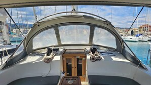 Interior de un barco con timón, dos asientos acolchados, una mesa de madera y vista al puerto deportivo con otros barcos y edificios.