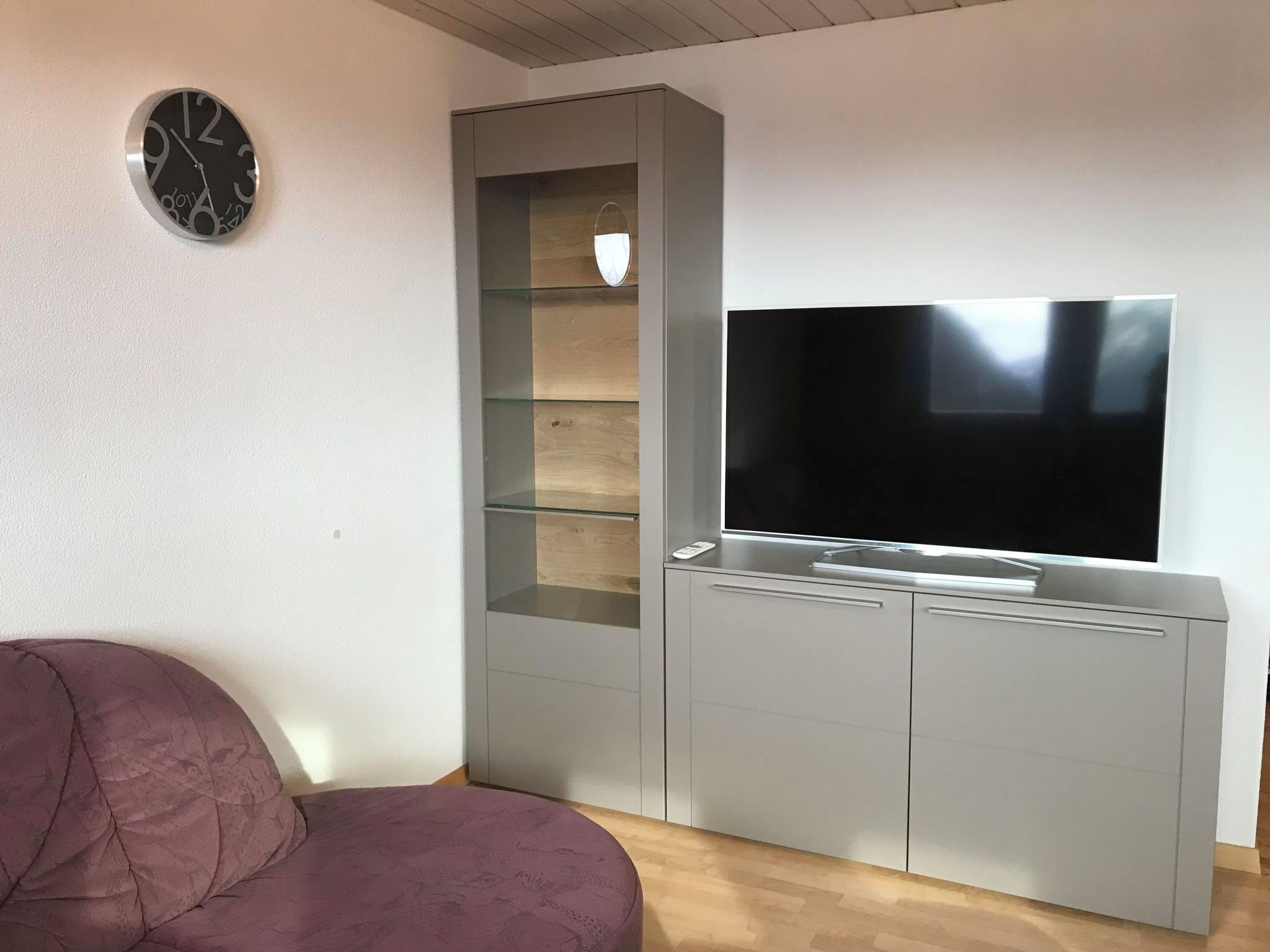 Un salon avec un meuble de rangement gris pour l’équipement audiovisuel, un téléviseur à écran plat et une horloge ronde fixée au mur.