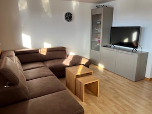 Una sala de estar moderna con un sofá seccional, una mesa de centro de madera, un televisor sobre un mueble bajo y un aparador de vidrio con estantes.