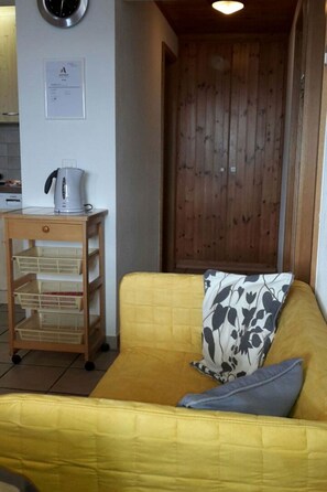 A yellow sofa with a patterned pillow and a grey pillow, a white kettle on a wooden side table, and a wooden door in the background.