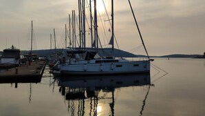A marina with multiple sailboats docked, calm water, and a distant hilly landscape.