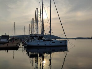 A marina with multiple sailboats docked, calm water, and a distant hilly landscape.
