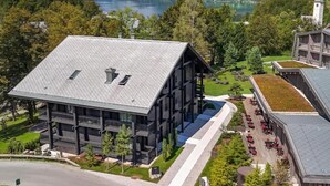 Un edificio moderno con techo inclinado, un patio con mesas al aire libre y vistas a un lago y montañas al fondo.
