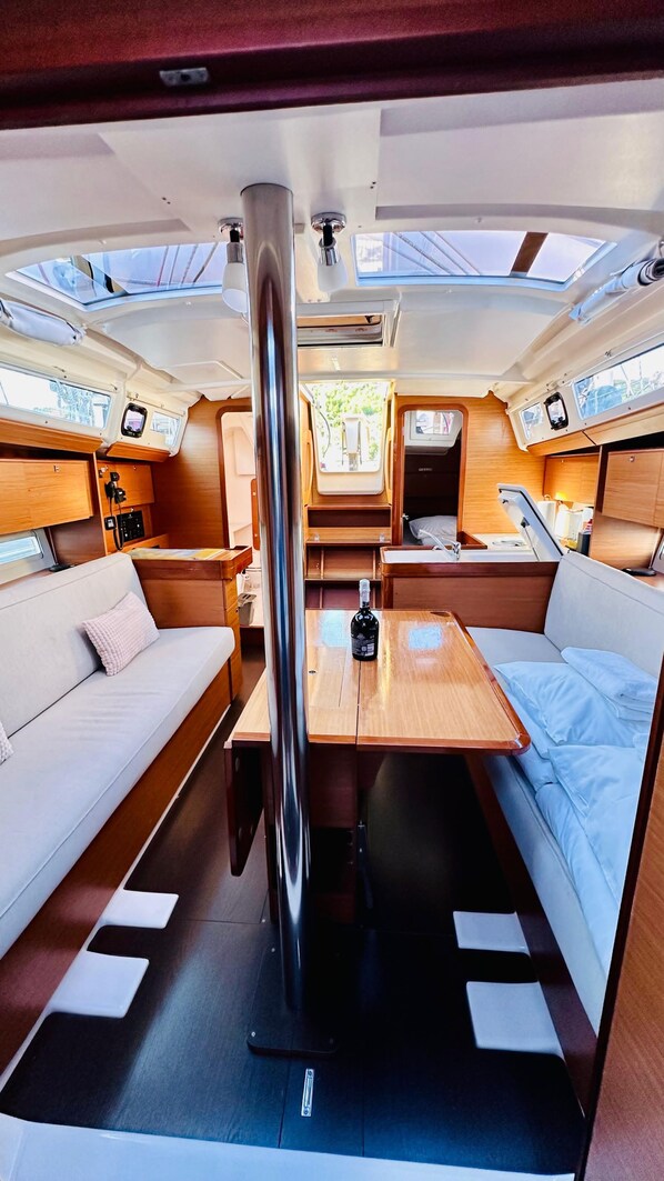 A boat interior with a central table, two sofas, a small kitchen area, and a sleeping berth.
