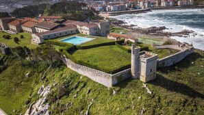 A coastal property with a swimming pool, a fortified structure, and a view of the sea and town.