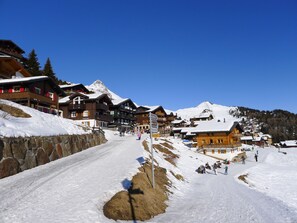 A snow-covered mountain village with wooden buildings, a stone wall, and a clear blue sky.
